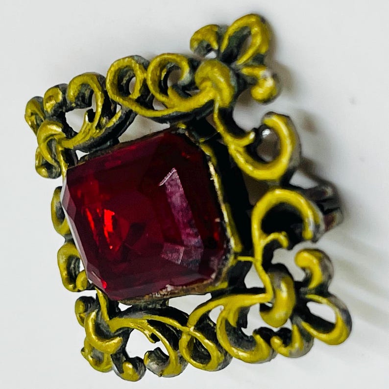 May include: A square brooch featuring a central faceted deep red gemstone set within a decorative frame of yellow-painted metal with intricate scrollwork patterns. The metal frame displays a dark, weathered finish beneath the yellow paint.