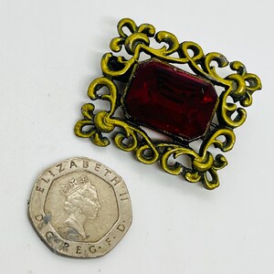 May include: A rectangular brooch featuring an ornate, dark gold-toned metal frame with scrollwork details surrounding a large, faceted deep red gemstone. The brooch is positioned next to a British twenty pence coin for scale reference.