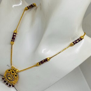 May include: A gold necklace with a pendant featuring a circular design with red stones. The necklace has a chain with small, round links and is adorned with burgundy-coloured beads.