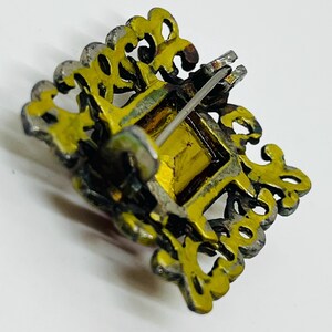 May include: A square brooch featuring a central amber-coloured stone set within a decorative, openwork gold-tone metal frame. The back of the piece displays a thin metal pin mechanism with a hinge and a clasp for securing the item to fabric.