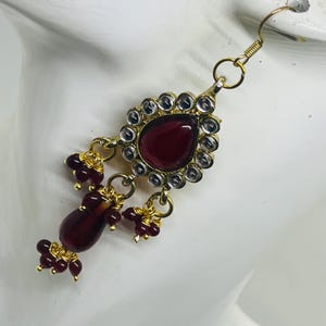 May include: A gold-tone dangle earring featuring a central teardrop-shaped deep red stone surrounded by small clear stones. The earring includes additional dangling deep red beads and smaller clusters of red beads attached by gold wire links.