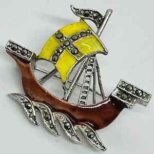 May include: A silver-toned brooch shaped like a sailing ship. The hull is finished in a glossy brown enamel, while the sail features a bright yellow enamel cross. The ship is accented with small, dark, faceted marcasite stones along the hull and sail.