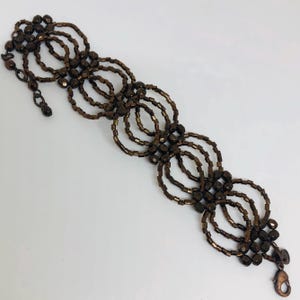 May include: A brown beaded bracelet with a woven design featuring multiple circles. The bracelet has a clasp closure.