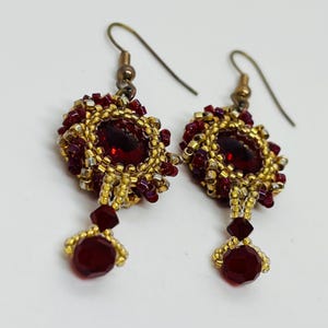 May include: A pair of handmade earrings featuring a central, faceted, deep red gemstone surrounded by gold and burgundy beads. The earrings have a teardrop design with a smaller red bead at the bottom and a gold-colored hook.