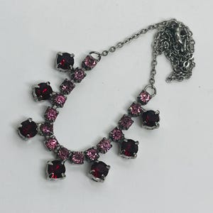 May include: A silver necklace with a fine chain. The necklace is adorned with square-cut, deep red and pink gemstones. The gemstones are set in silver metal. The necklace is presented on a white surface.