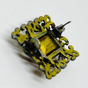 May include: A square brooch featuring a central amber-coloured gemstone set within a dark, ornate metal frame. The frame has an intricate, openwork filigree design with scalloped edges. A metal pin mechanism is visible on the back of the piece.