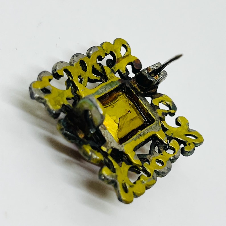 May include: A square brooch featuring a central amber-coloured gemstone set within a dark, ornate metal frame. The frame has an intricate, openwork filigree design with scalloped edges. A metal pin mechanism is visible on the back of the piece.