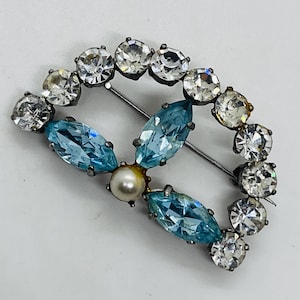 May include: Oval-shaped vintage brooch with clear and light blue crystals. The brooch features a central design of three light blue marquise-cut crystals and a pearl, surrounded by clear round crystals. The pin is silver-toned.