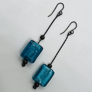 May include: A pair of dangle earrings featuring rectangular, teal-colored glass beads. The beads are suspended from dark metal wires with small, round beads and curved ear wires. The earrings have a simple, elegant design.