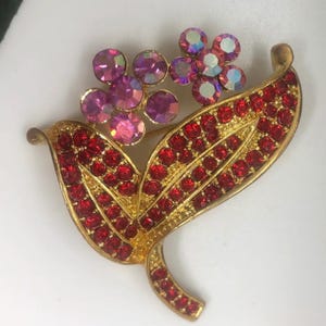 May include: Gold tone brooch with red and pink rhinestones. The brooch features a stylised flower design with leaves.