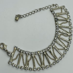 May include: Silver-toned bracelet with a decorative design. The bracelet features a series of vertical bars and small, round, clear gemstones. A chain extends from the bracelet for adjustable sizing, and a clasp is present for closure.