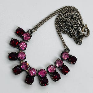 May include: A vintage necklace featuring a series of alternating pink and red gemstones set in a silver-tone metal. The necklace has a delicate chain and a clasp closure. The gemstones are oval and round, creating a symmetrical design.