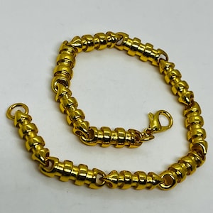 May include: A gold-colored bracelet with a unique design. The bracelet features a series of connected, cylindrical links, creating a textured appearance. It has a lobster clasp closure for secure wear. The jewelry is displayed on a white surface.