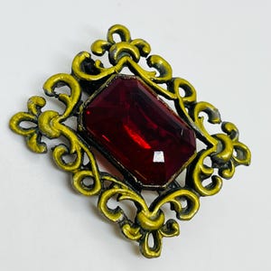 May include: A rectangular brooch featuring a large, faceted deep red gemstone set within an ornate, scrollwork frame finished in a distressed, matte yellow-gold tone. The metal frame displays intricate, symmetrical curves and pointed fleur-de-lis accents.
