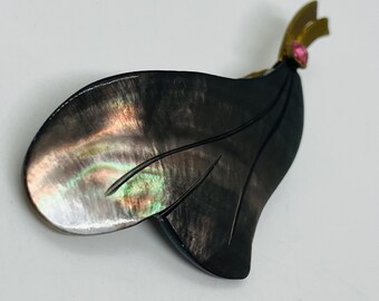 May include: A leaf-shaped brooch with a dark, iridescent surface, featuring shades of black, green, and pink. The brooch has a gold-colored accent and a small pink gemstone. The design is elegant and nature-inspired.