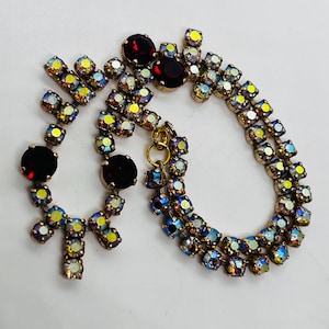 May include: Vintage gold-toned necklace and earring set. The necklace and earrings feature a combination of iridescent, multi-coloured rhinestones and large, dark red stones. The rhinestones are set in a gold-toned metal setting.