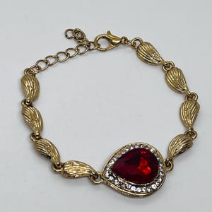 May include: Gold-toned bracelet featuring a teardrop-shaped, red gemstone centerpiece surrounded by small, clear crystals. The bracelet has a series of leaf-shaped links and an adjustable clasp.