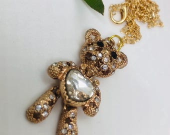 May include: Gold-tone necklace featuring a teddy bear pendant embellished with clear and black rhinestones. The bear is holding a clear heart-shaped crystal.