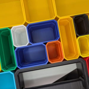 May include: An assortment of colorful plastic storage bins in various sizes and shapes. The bins are in shades of yellow, green, white, blue, orange, black, and red. The bins are arranged in a grid-like pattern, with some overlapping.