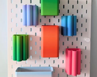 Könnte beinhalten: Eine Sammlung farbenfroher, geometrischer Aufbewahrungsbehälter in verschiedenen Formen und Größen, die an einer weißen Lochwand befestigt sind. Die Behälter sind in Grün-, Blau-, Orange- und Rosatönen gehalten und bieten eine lebendige und organisierte Präsentation.