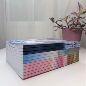 Könnte beinhalten: Ein Stapel Notizbücher mit verschiedenen farbigen Umschlägen. Die Umschläge zeigen eine Vielzahl von abstrakten Designs, darunter blau und weiß, rosa und gold sowie ein blaues und weißes Aquarelldesign.