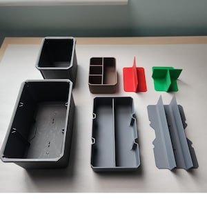 May include: A collection of modular storage components including two dark gray rectangular bins, a brown multi-compartment tray, a gray divided tray, a red T-shaped divider, a green cross-shaped divider, and a gray H-shaped structural insert.