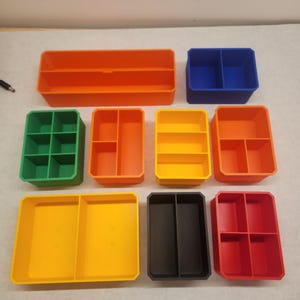 May include: A collection of colorful, plastic storage containers in various shapes and sizes. The containers are in shades of orange, blue, green, yellow, black, and red. Some have multiple compartments, while others are open trays. Ideal for organizing small items.