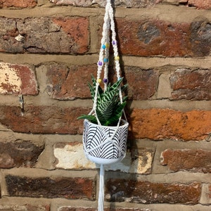 Peut inclure: Un porte-plante en macramé blanc avec un pot blanc et une plante succulente verte. Le porte-plante est orné de perles colorées dans les tons jaune, bleu et violet.