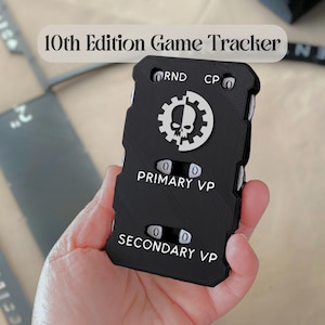 May include: A black 10th Edition Game Tracker with a white skull and gear emblem. The device features mechanical dials for RND, CP, PRIMARY VP, and SECONDARY VP tracking. The rectangular unit has a textured surface and white printed text labels.