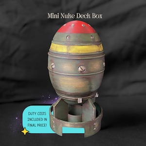 May include: A novelty Mini Nuke Deck Box, painted in olive green, yellow, and red. The box is shaped like a bomb with a rounded top and a base with fins. The text "Mini Nuke Deck Box" is at the top.