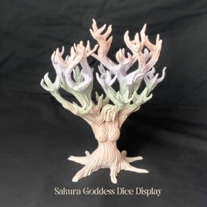 May include: A white, pink and green ceramic tree sculpture with a female face in the trunk. The tree has many branches that reach upwards and are shaped like hands. The sculpture is labelled "Sakura Goddess Dice Display".