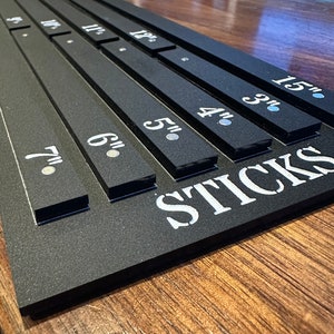 Puede incluir: Porta-palos de madera negro con medidas en pulgadas de 7 a 12 pulgadas. El porta-palos tiene la palabra "STICKS" impresa en la parte inferior.