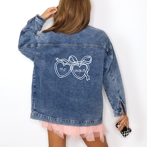 May include: A light-wash denim jacket with white embroidered hearts and the text "H-S 09.06.23". The jacket has long sleeves and a classic collar. The model is wearing a pink tulle skirt and white boots.