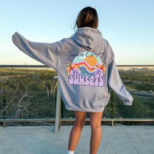 May include: A grey hooded sweatshirt with a graphic print on the back. The graphic features a colourful sunset with the text "Forever Chasing Sunsets".