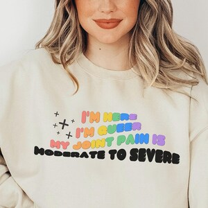 Könnte beinhalten: Ein hellbeiger Pullover mit dem Text "I'm here I'm queer My joint pain is moderate to severe" in Regenbogenfarben.