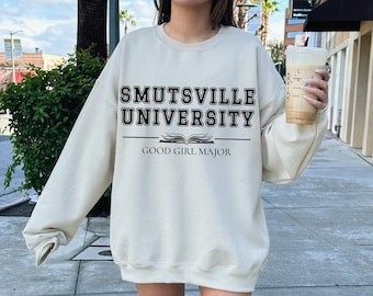May include: A cream-colored sweatshirt with the text "Smutsville University" and "Good Girl Major" printed in black. The sweatshirt is worn by a person standing on a sidewalk.