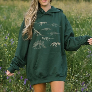 Puede incluir: Una sudadera con capucha verde oscuro con un estampado gráfico de esqueletos de dinosaurios. La sudadera tiene un bolsillo canguro y una capucha con cordón.
