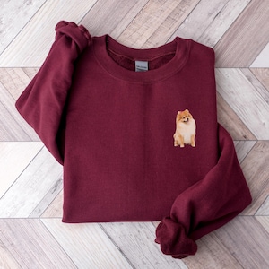 Könnte beinhalten: Ein maroonfarbener Pullover mit einem kleinen gestickten Bild eines braunen und weißen Zwergspitz-Hundes auf der linken Brust.