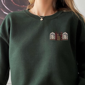 May include: A dark green sweatshirt with a small embroidered design on the front. The design features two stylised buildings with a red, white and blue colour scheme.