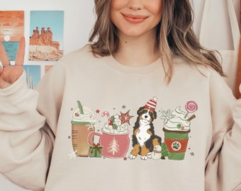 Puede incluir: Una sudadera beige con un diseño navideño que presenta un perro de montaña bernés sentado frente a una fila de tazas de café con temática navideña. Las tazas están decoradas con crema batida, bastones de caramelo y copos de nieve. El perro lleva un gorro de Papá Noel.