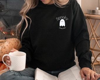 May include: Black sweatshirt with a white ghost graphic and the words "Spooky Season". The sweatshirt is paired with blue jeans. A white mug is held in the hand. A book is open in the lap.