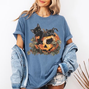 Peut inclure: Un t-shirt bleu avec un motif de trois chatons noirs &agrave; l'int&eacute;rieur d'une citrouille. Les chatons regardent hors de la citrouille, qui a un visage sculpt&eacute;. La citrouille est entour&eacute;e de feuilles vertes.