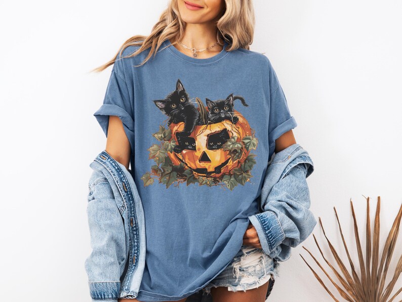 Peut inclure: Un t-shirt bleu avec un motif de trois chatons noirs &agrave; l'int&eacute;rieur d'une citrouille. Les chatons regardent hors de la citrouille, qui a un visage sculpt&eacute;. La citrouille est entour&eacute;e de feuilles vertes.