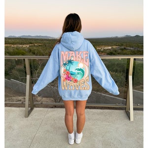May include: Light blue hoodie with the words "MAKE WAVES" in a retro design on the back. The graphic includes a tropical scene with palm trees, a wave, and flowers. The hoodie is worn by a person.