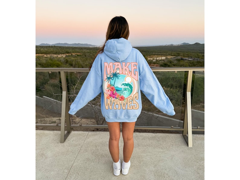 May include: Light blue hoodie with the words "MAKE WAVES" in a retro design on the back. The graphic includes a tropical scene with palm trees, a wave, and flowers. The hoodie is worn by a person.