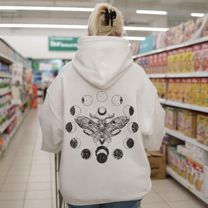 May include: White hoodie with a black moth and moon phase design on the back. The graphic features a detailed moth illustration surrounded by circular moon phase symbols. The hoodie has a hood and long sleeves, and is made of a soft material.