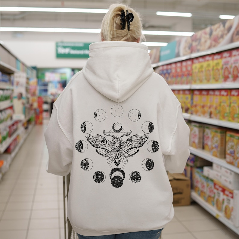 May include: White hoodie with a black moth and moon phase design on the back. The graphic features a detailed moth illustration surrounded by circular moon phase symbols. The hoodie has a hood and long sleeves, and is made of a soft material.