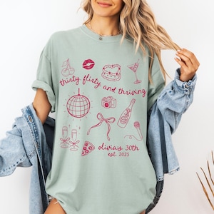 Może przedstawiać: Jasnozielony t-shirt z r&oacute;żowym nadrukiem graficznym przedstawiającym kulę dyskotekową, butelkę szampana, wiśnie, kawałek pizzy, aparat fotograficzny, szminkę, ciasto i kieliszek martini. Tekst na koszulce brzmi "thirty flirty and thriving Olivia's 30th est. 2025".