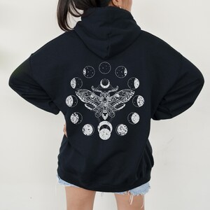 May include: Black hooded sweatshirt with a white graphic of a moth and phases of the moon. The moth design is centered on the back of the sweatshirt. The moon phases are arranged in a circle around the moth.
