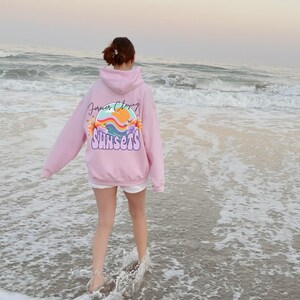 May include: A pink hoodie with the text "Forever Chasing Sunsets" and a graphic of a sunset with flowers on the back. The person wearing the hoodie is walking on a sandy beach with waves.